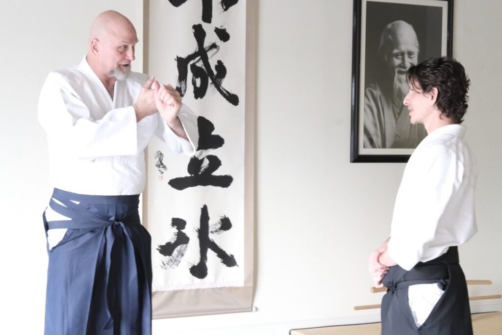 Ellis Amdur on the Dojo’s Second Promise – Aikido Journal Ellis Amdur, in hakama, explains a point to Josh Gold beneath calligraphy and a portrait of the Founder.